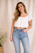 Woman wearing a white top and blue jeans standing against a beige curtain.