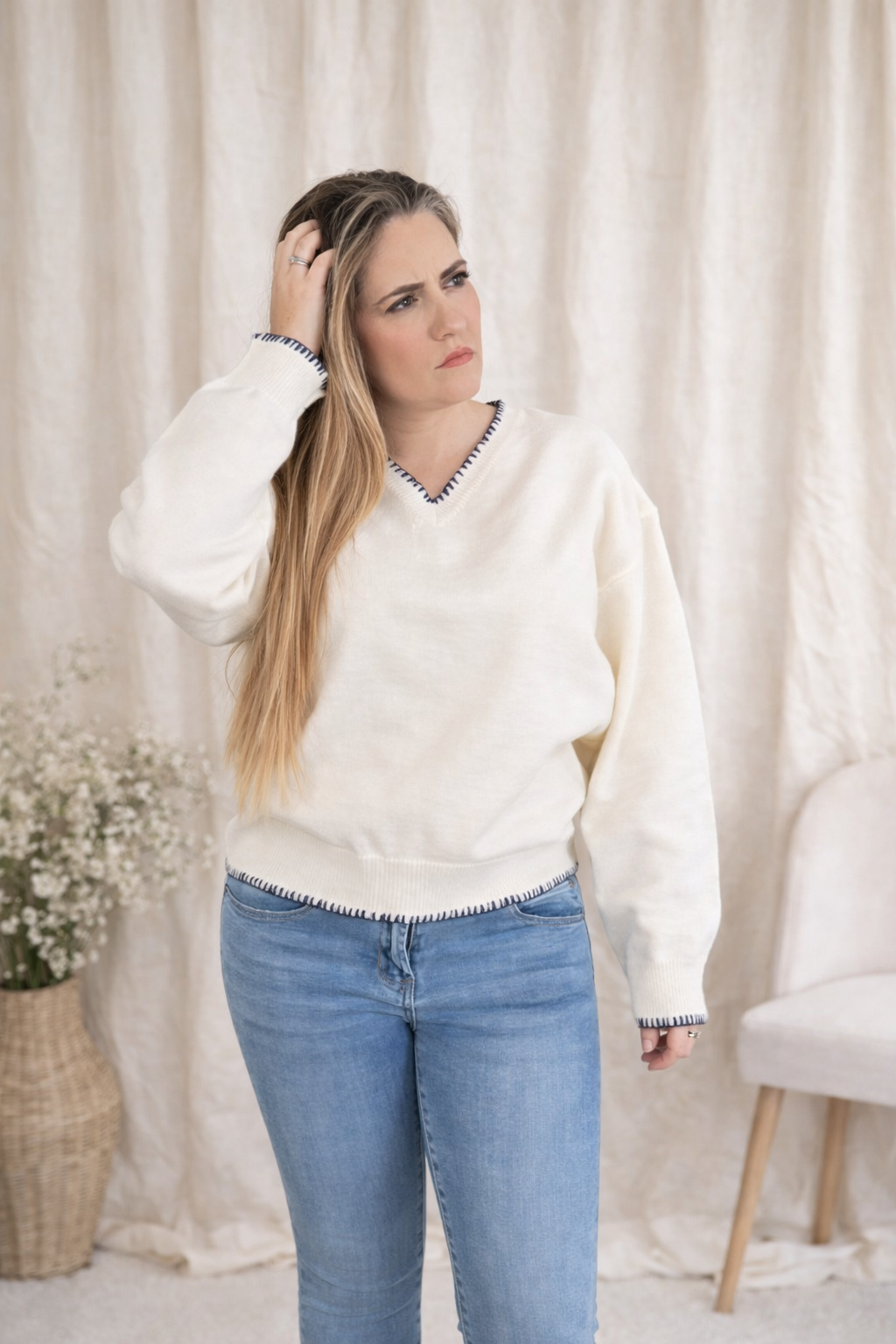 Woman wearing a white sweater and blue jeans standing against a white curtain.