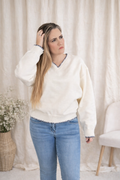 Woman wearing a white sweater and blue jeans standing against a white curtain.