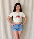Young girl wearing a white t-shirt with a graphic design and denim shorts against a beige curtain background