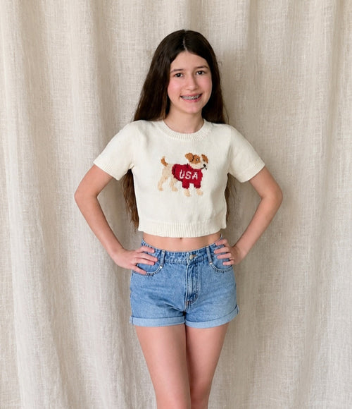 Young girl wearing a white t-shirt with a graphic design and denim shorts against a beige curtain background