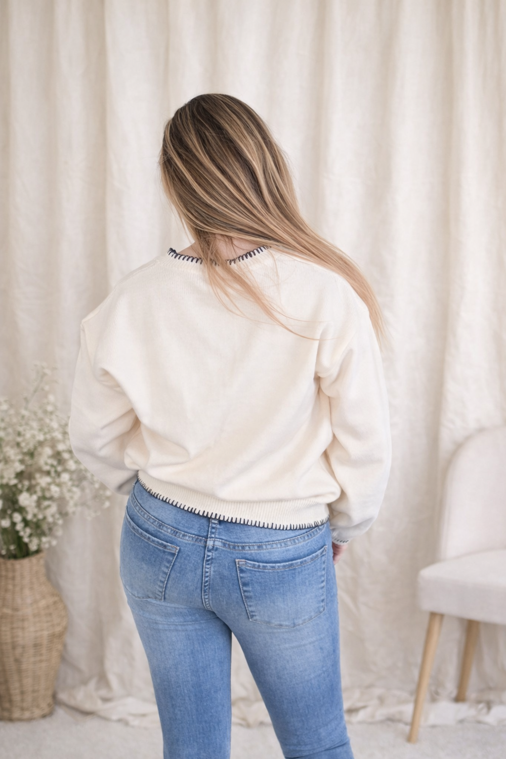 Person wearing a white sweater and blue jeans standing in front of a white curtain.