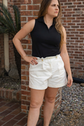 Woman in black sleeveless top and white shorts standing against a brick wall.