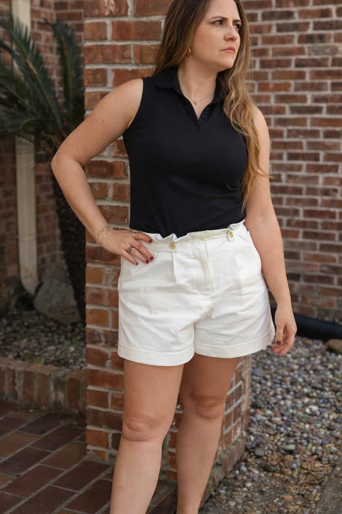 Woman in black sleeveless top and white shorts standing against a brick wall.
