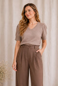 A woman wearing a taupe short-sleeve V-neck knit top and brown high-waisted wide-leg trousers, standing against a neutral beige curtain backdrop.