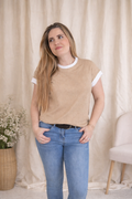 Woman wearing a beige sweater and blue jeans standing against a beige curtain.